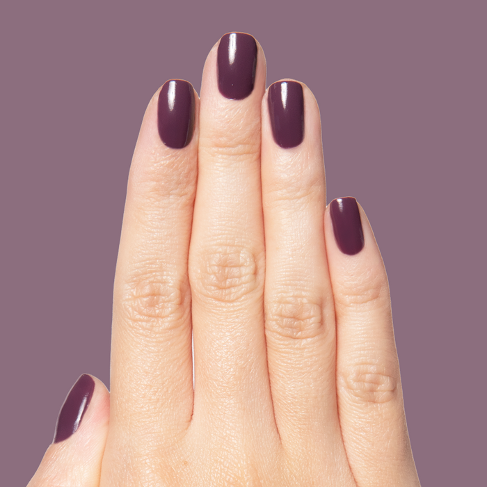 Hand with nails painted in glossy Plum gel polish showing a smooth quick dry manicure on a muted purple background