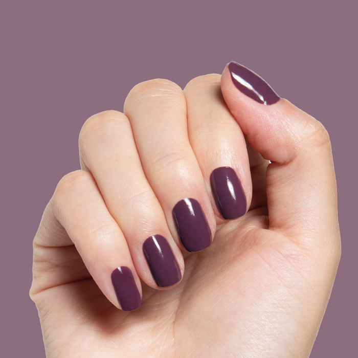 Hand with nails painted in glossy Plum gel polish showing a smooth quick dry manicure on muted purple background