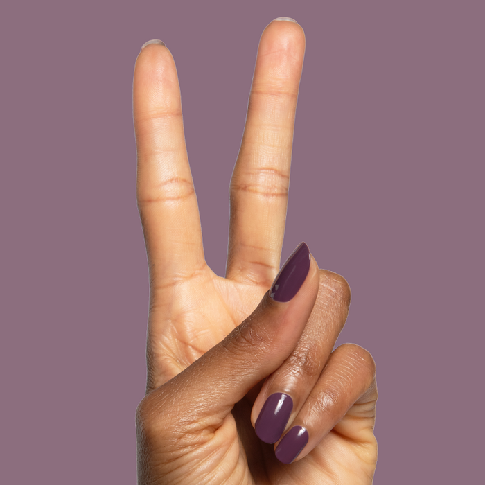Hand with Plum polished nails showing a glossy quick dry gel manicure on a muted purple background