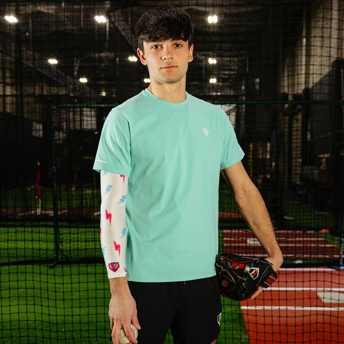 Male baseball player in a mint green shirt wearing the lightning arm sleeve on his left arm while holding a glove and baseball in an indoor batting cage