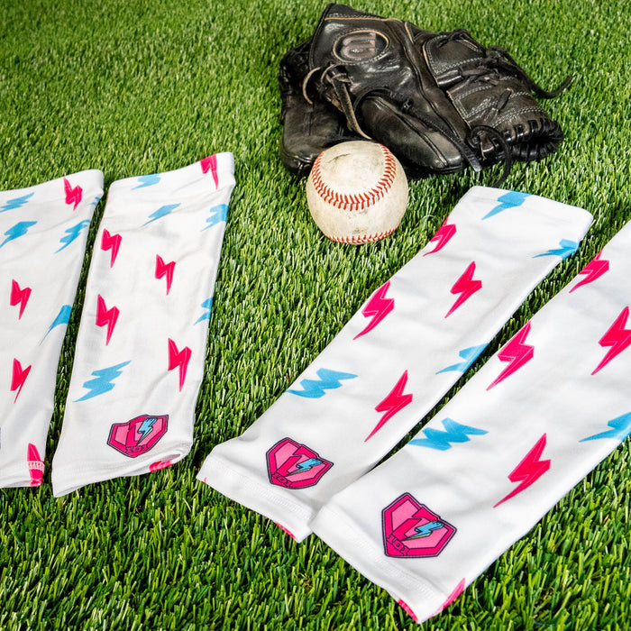 Two lightning arm sleeves laid on green turf next to a baseball and a glove showing the pink and blue lightning bolt print