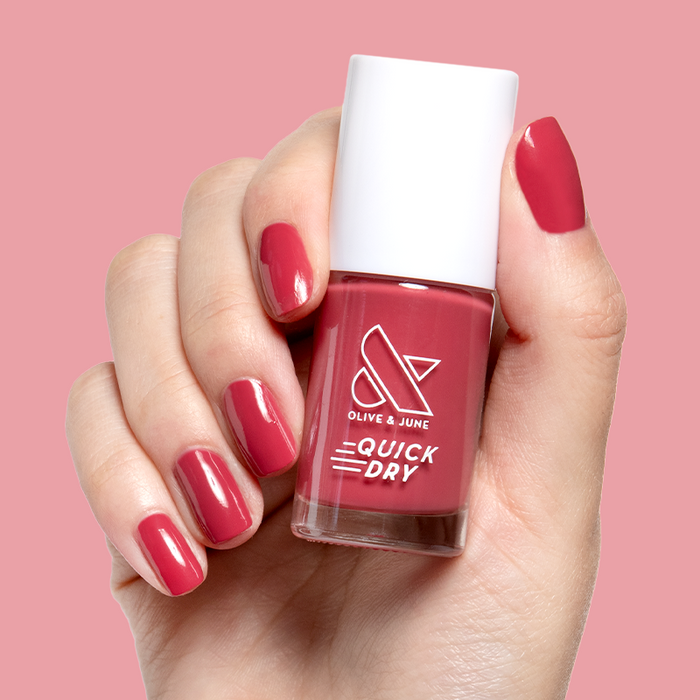 Hand with natural nails painted in bold pinky red Roar quick dry gel polish holding bottle against matching background