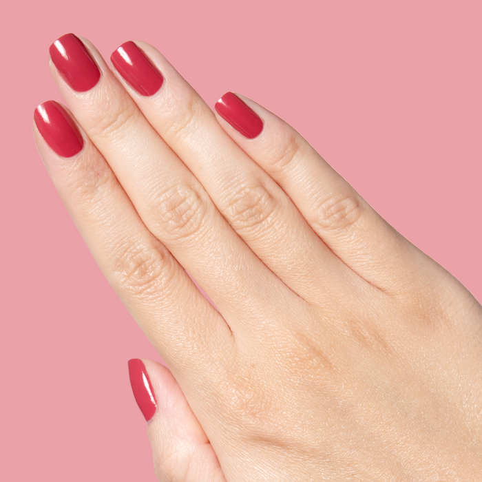 Hand with neatly shaped nails painted in bold pinky red Roar gel polish against a soft pink background