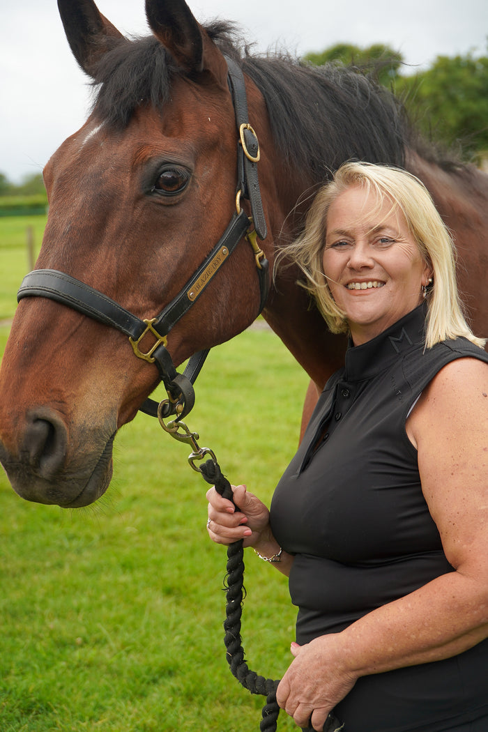 Woman standing next to a horse in an outdoor setting.  Ultra-soft technical sport fabric. Breathable and lightweight for warmer weather. Flattering athletic fit
Signature black branded Mayston collar. Subtle gloss shoulder logo detail. Sleeveless design for unrestricted movement. blackout colour match branding