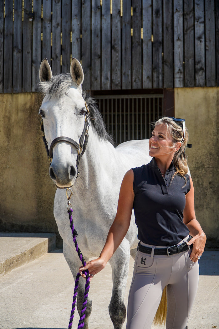 Woman in equestrian attire standing next to a white horse in an outdoor setting.  Ultra-soft technical sport fabric. Breathable and lightweight for warmer weather. Flattering athletic fit
Signature black branded Mayston collar. Subtle gloss shoulder logo detail. Sleeveless design for unrestricted movement. blackout colour match branding