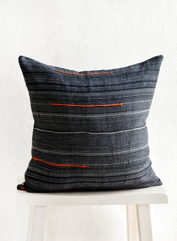 A square throw pillow made from dark blue fabric with grey stripes and embroidered orange lines.