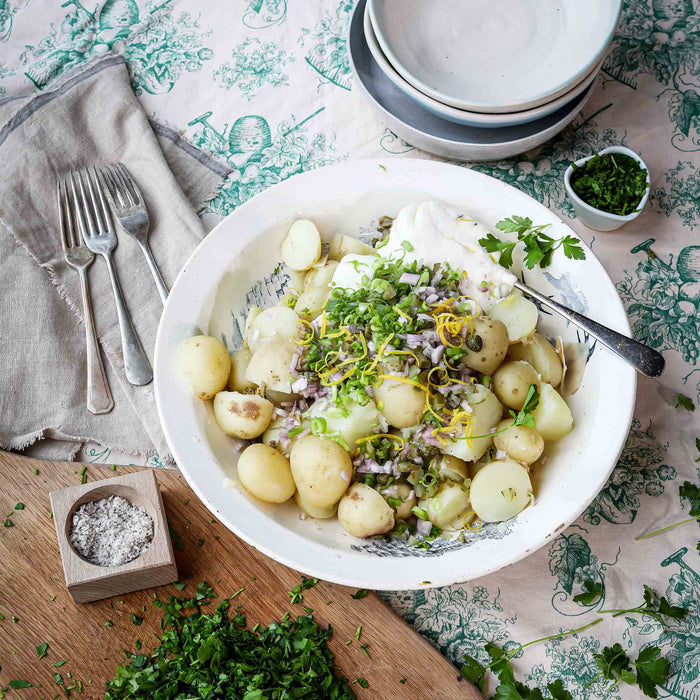 Boiled chopped 'swift' potatoes with fresh herbs and mayo on top for making a potato salad. Sat in a bowl on a table with table cloth and cutlery.