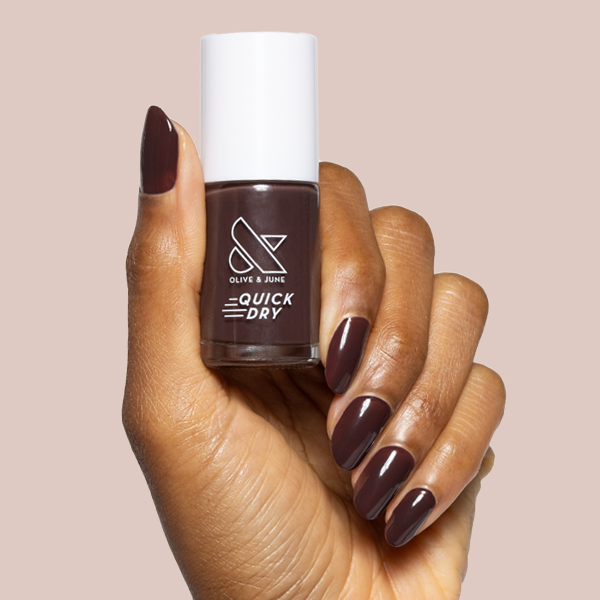Hand holding Spressotini quick dry gel polish bottle with dark coffee brown manicure on medium nails against soft beige background