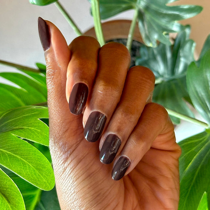 Hand with shiny Spressotini gel polish manicure in deep coffee brown on medium-length nails against green foliage background
