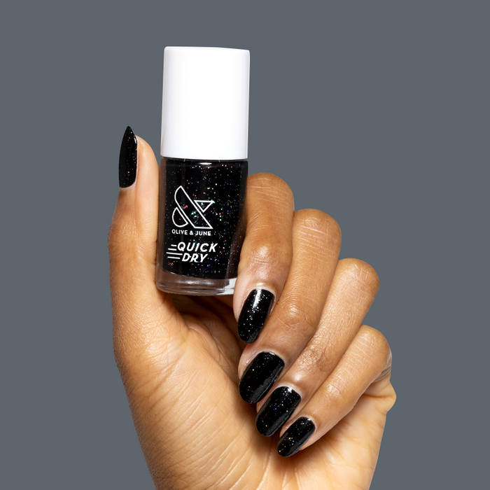 Hand holding Olive and June Stargaze quick dry gel polish bottle with matching glossy black nails and rainbow glitter polish