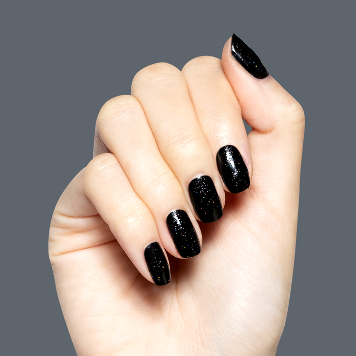 Hand with Stargaze gel polish manicure in glossy black with fine rainbow glitter on medium-length natural nails against gray background