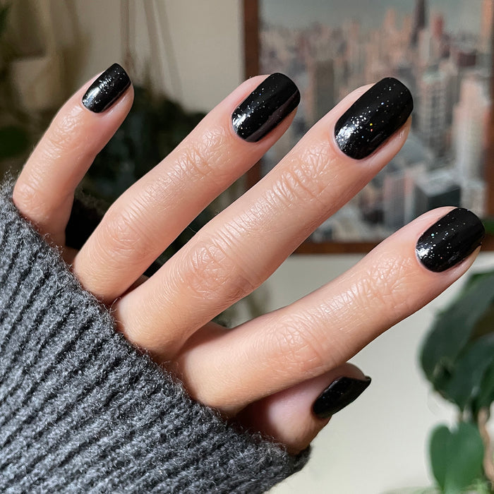 Hand with Stargaze gel polish manicure in glossy black speckled with tiny rainbow glitter on medium-length nails against blurred background
