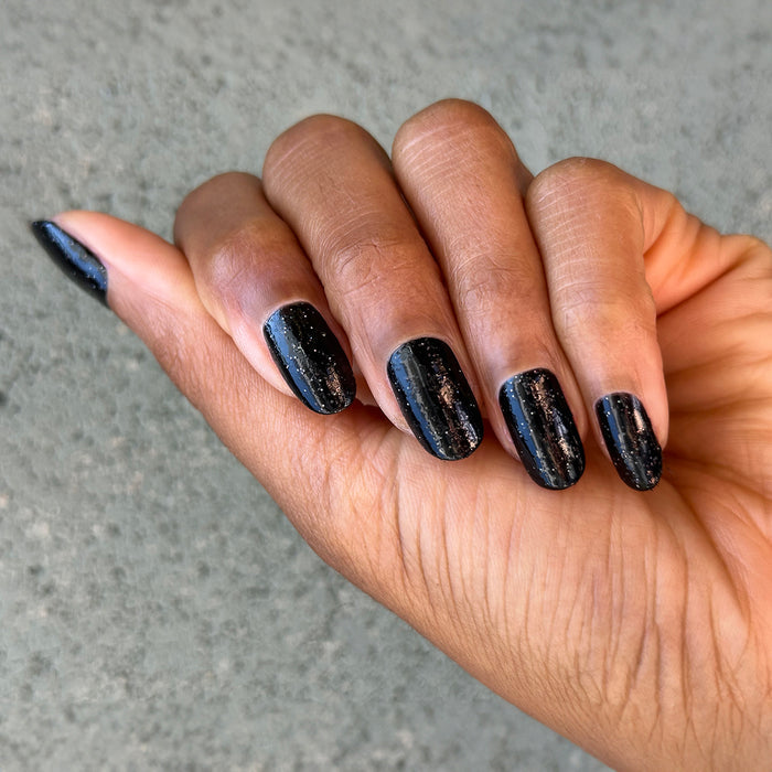 Hand with Stargaze gel polish manicure in glossy deep black with shimmering rainbow glitter on medium-length nails against gray surface