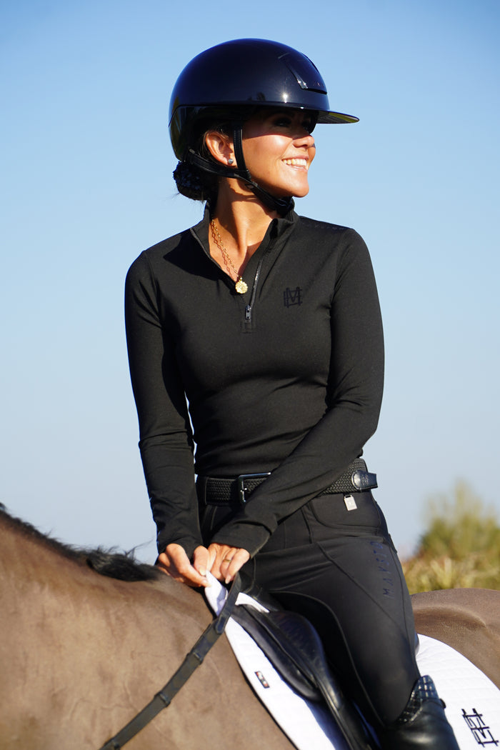 Person riding a horse with a clear blue sky background. Sleek silhouette baselayer top subtle Mayston branding, maximum coverage without compromising style,  next-level performance with a bold, minimalist look. Moisture-wicking, breathable fabric with four-way stretch, it keeps you cool and comfortable. Training, Everyday or competition wear. longsleeve 1/4 zip with collar. black with black branding
