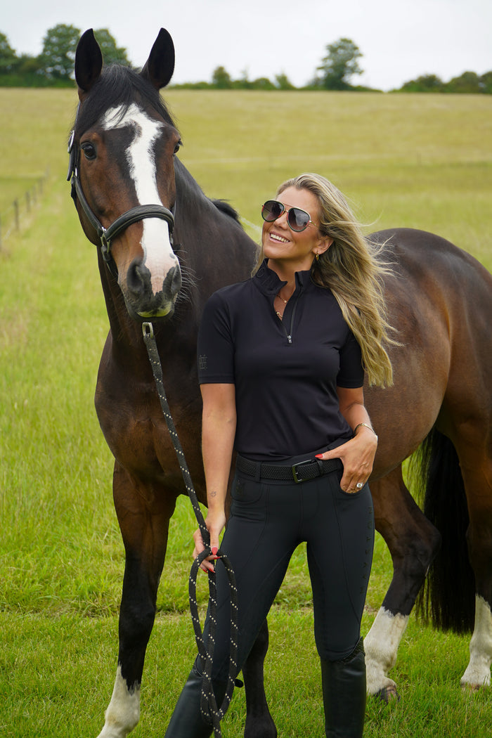 Woman standing next to a horse in a grassy field. Broadstone Short sleeve Luxury Sports material base layer short sleeve. Black with subtle black branding colour match embroidery. High collar that's not restrictive. 1/4 zip. Blackout design