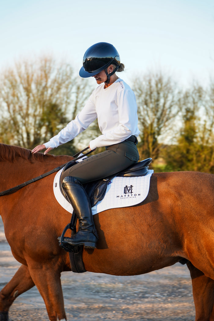 Person riding a horse with a saddle pad featuring a logo, set against a natural background. Premium Dark Green breeches with a Full Silicone seat. Stretch high waisted Jodhpurs. Deep pockets, supportive, comfortable and compression quality fabric. Elegant detailing with subtle branding. Hunter Green