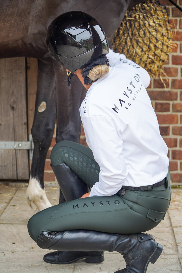 Person in equestrian attire with a horse, wearing a white jacket and green riding pants, in front of a brick wall. Premium Dark Green breeches with a Full Silicone seat. Stretch high waisted Jodhpurs. Deep pockets, supportive, comfortable and compression quality fabric. Elegant detailing with subtle branding. Hunter Green