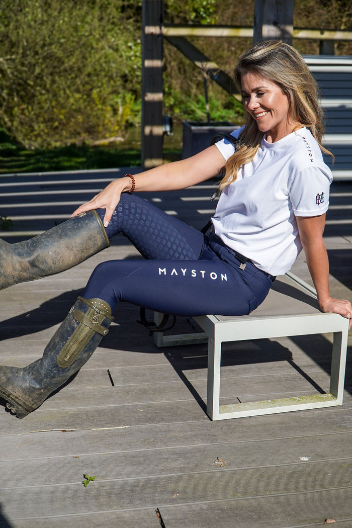 Woman sitting on a bench wearing navy leggings with 'Mayston' branding and a white shirt.  Premium navy breeches with a Full Silicone seat. Stretch high waisted Jodhpurs. Deep pockets, supportive, comfortable and compression quality fabric. Elegant detailing with subtle branding. high gloss white branding