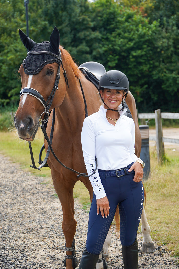 Woman in equestrian attire standing next to a brown horse on a gravel path with greenery in the background.  Premium navy breeches with a Full Silicone seat. Stretch high waisted Jodhpurs. Deep pockets, supportive, comfortable and compression quality fabric. Elegant detailing with subtle branding. high gloss white branding