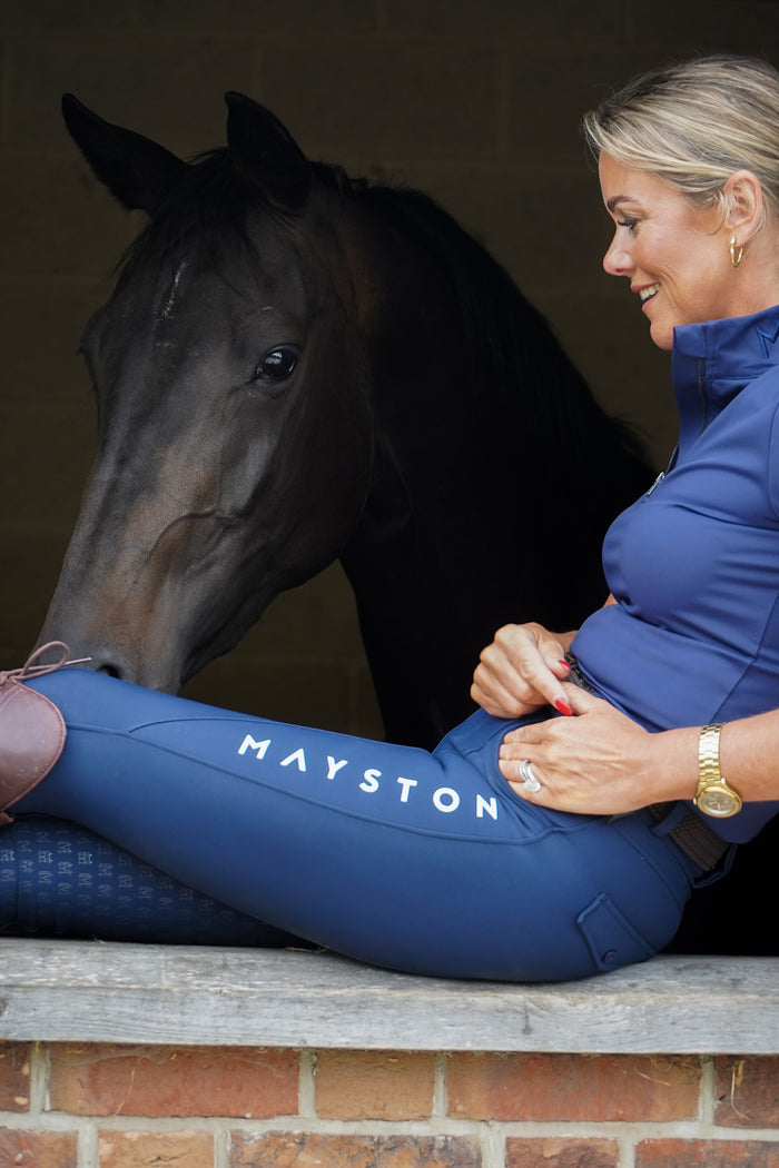 Woman in blue equestrian attire sitting next to a horse with 'Mayston'  Premium navy breeches with a Full Silicone seat. Stretch high waisted Jodhpurs. Deep pockets, supportive, comfortable and compression quality fabric. Elegant detailing with subtle branding. high gloss white brandingbranding.