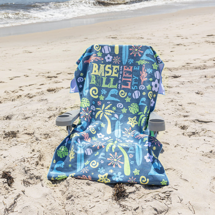 A beach chair with a colorful 'BASEBALL LIFESTYLE' print sits on a sandy beach with the ocean waves in the background.