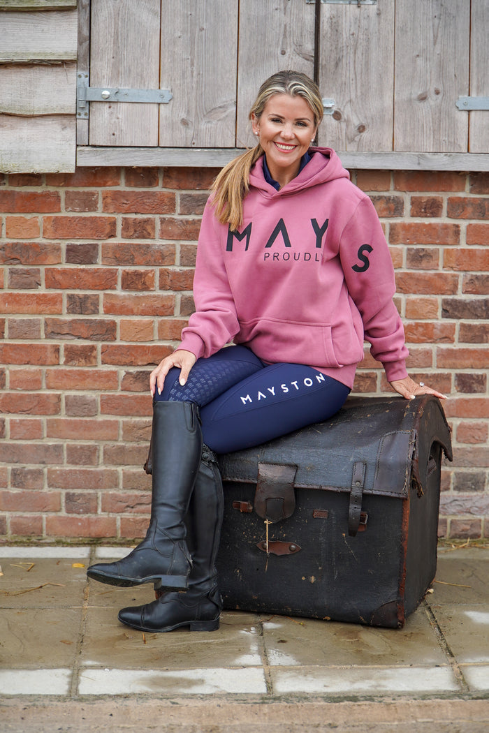 Woman in pink hoodie and blue breeches sitting on a vintage suitcase with a brick wall background. High quality luxurious 100% cotton. wrap around bold logo. soft, cozy and  warm. Hoodie, jumper sweatshirt. everyday wear. casual and smart