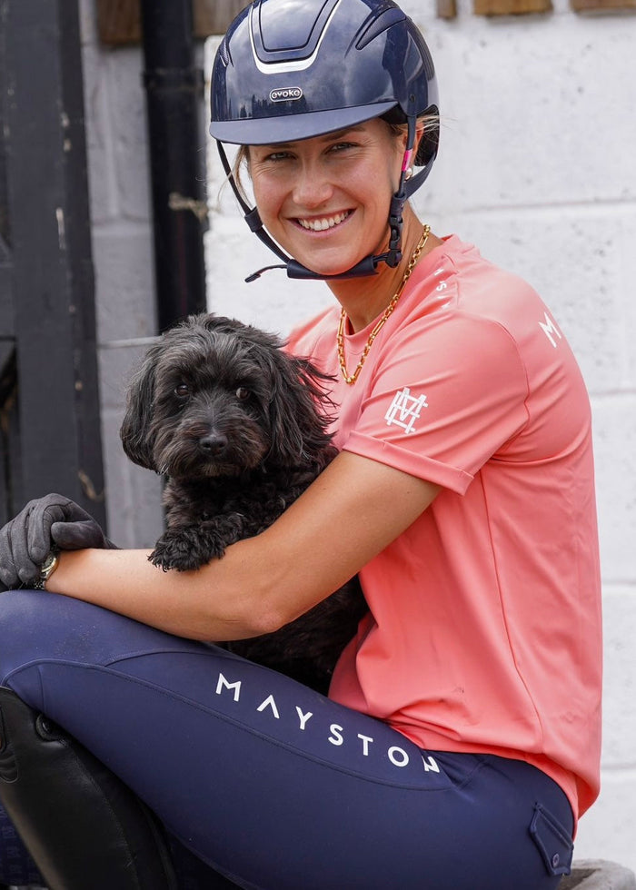 A person wearing a coral pink long-sleeved equestrian t-shirt holding a black dog luxury quality t shirt and fabric is specially crafted for active duty, providing flexibility and ease of movement for any busy lifestyle. An ideal choice for a day at the yard, in the saddle, or during your grooming duties, its loose-fitting design guarantees complete freedom.