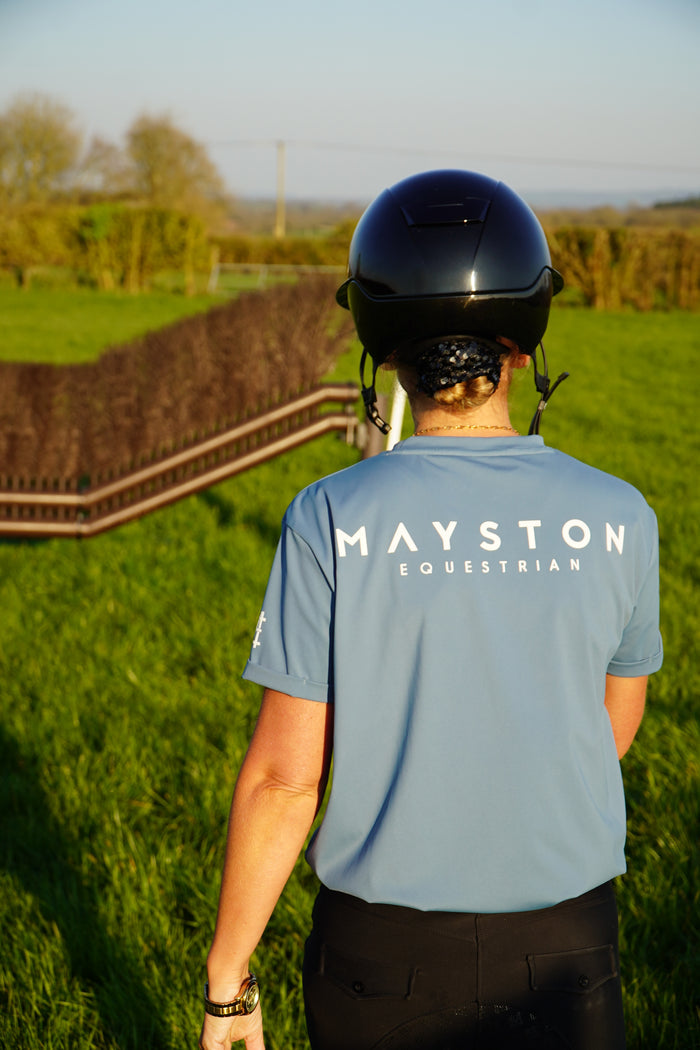 Person wearing a blue shirt with 'MAYSTON' branding, standing on grass with a path and trees in the background. A person wearing a blue t-shirt with the text 'MAYSTON' on the back, standing in an outdoor setting. luxury quality t shirt and fabric is specially crafted for active duty, providing flexibility and ease of movement for any busy lifestyle. An ideal choice for a day at the yard, in the saddle, or during your grooming duties, its loose-fitting design guarantees complete freedom. high glo