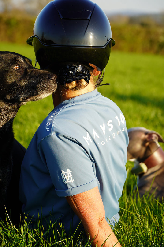 Person wearing a helmet and blue shirt with a dog on a grassy field. A person wearing a blue t-shirt with the text 'MAYSTON' on the back, standing in an outdoor setting. luxury quality t shirt and fabric is specially crafted for active duty, providing flexibility and ease of movement for any busy lifestyle. An ideal choice for a day at the yard, in the saddle, or during your grooming duties, its loose-fitting design guarantees complete freedom. high gloss white branding on a dorset blue colour