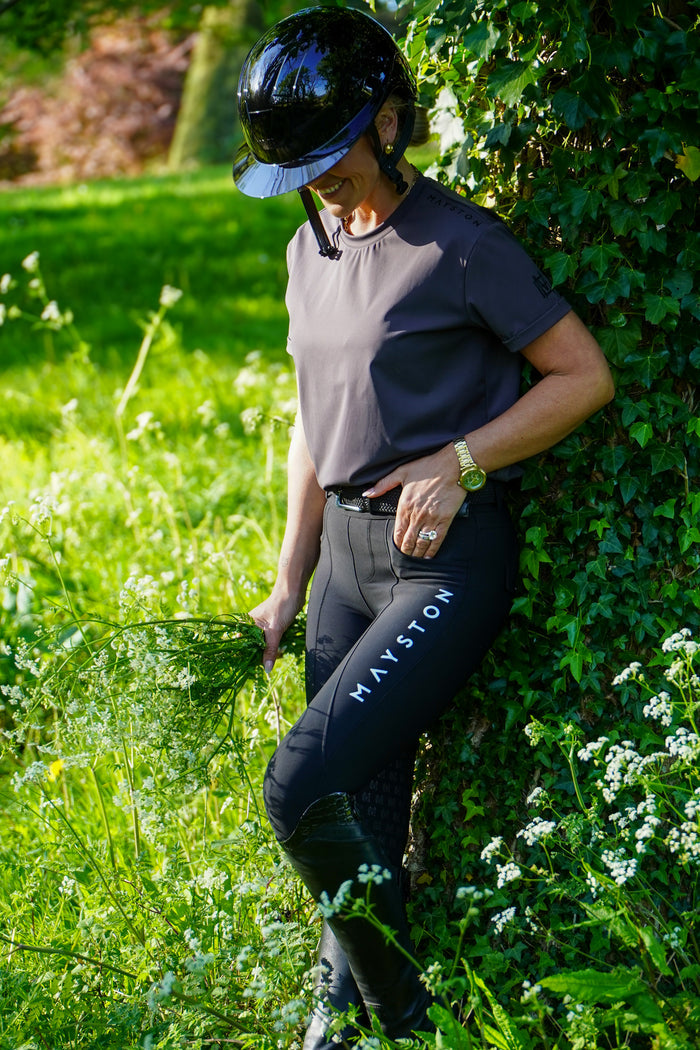 Person in equestrian attire standing in a grassy area with greenery. grey luxury quality t shirt and fabric is specially crafted for active duty, providing flexibility and ease of movement for any busy lifestyle. An ideal choice for a day at the yard, in the saddle, or during your grooming duties, its loose-fitting design guarantees complete freedom. high gloss black silicone branding