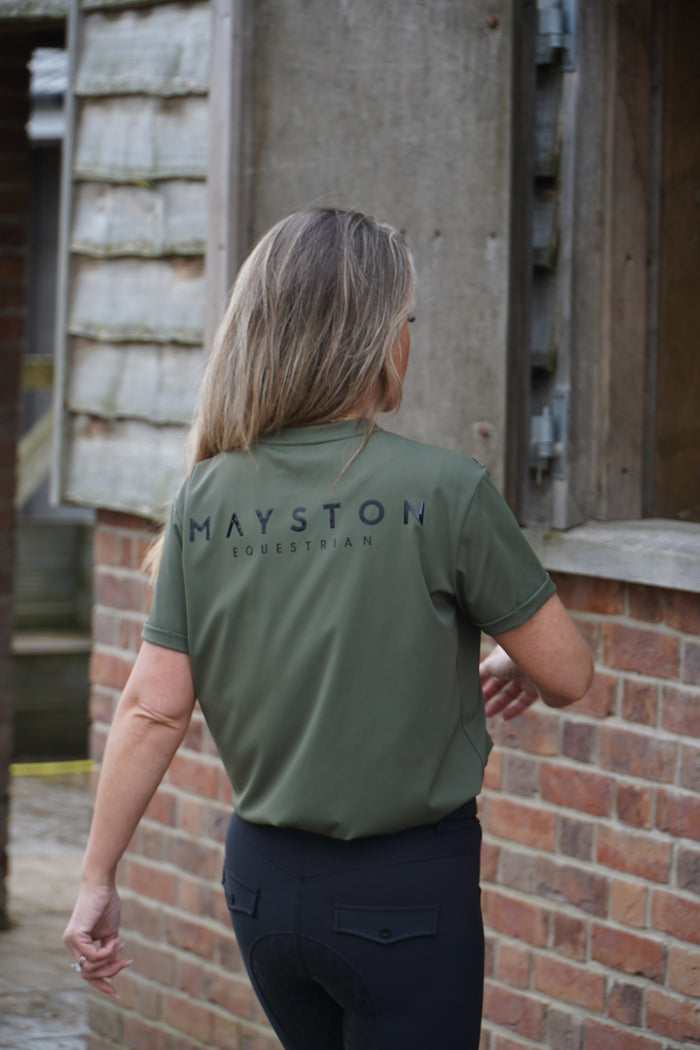 Person wearing a green shirt with 'Mayston' branding, standing in front of a brick wall. luxury quality t shirt and fabric is specially crafted for active duty, providing flexibility and ease of movement for any busy lifestyle. An ideal choice for a day at the yard, in the saddle, or during your grooming duties, its loose-fitting design guarantees complete freedom. Hunter green colour