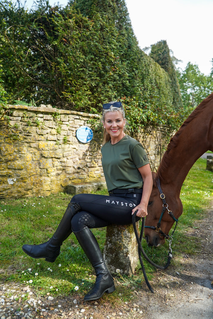Woman sitting on a stone bench next to a horse in a garden setting. luxury quality t shirt and fabric is specially crafted for active duty, providing flexibility and ease of movement for any busy lifestyle. An ideal choice for a day at the yard, in the saddle, or during your grooming duties, its loose-fitting design guarantees complete freedom. Hunter green colour