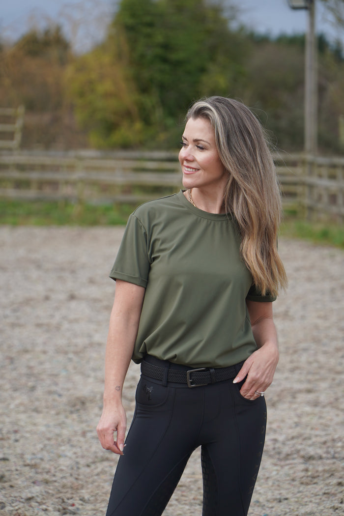 Woman in green shirt and black pants standing outdoors with a blurred background. luxury quality t shirt and fabric is specially crafted for active duty, providing flexibility and ease of movement for any busy lifestyle. An ideal choice for a day at the yard, in the saddle, or during your grooming duties, its loose-fitting design guarantees complete freedom. Hunter green colour