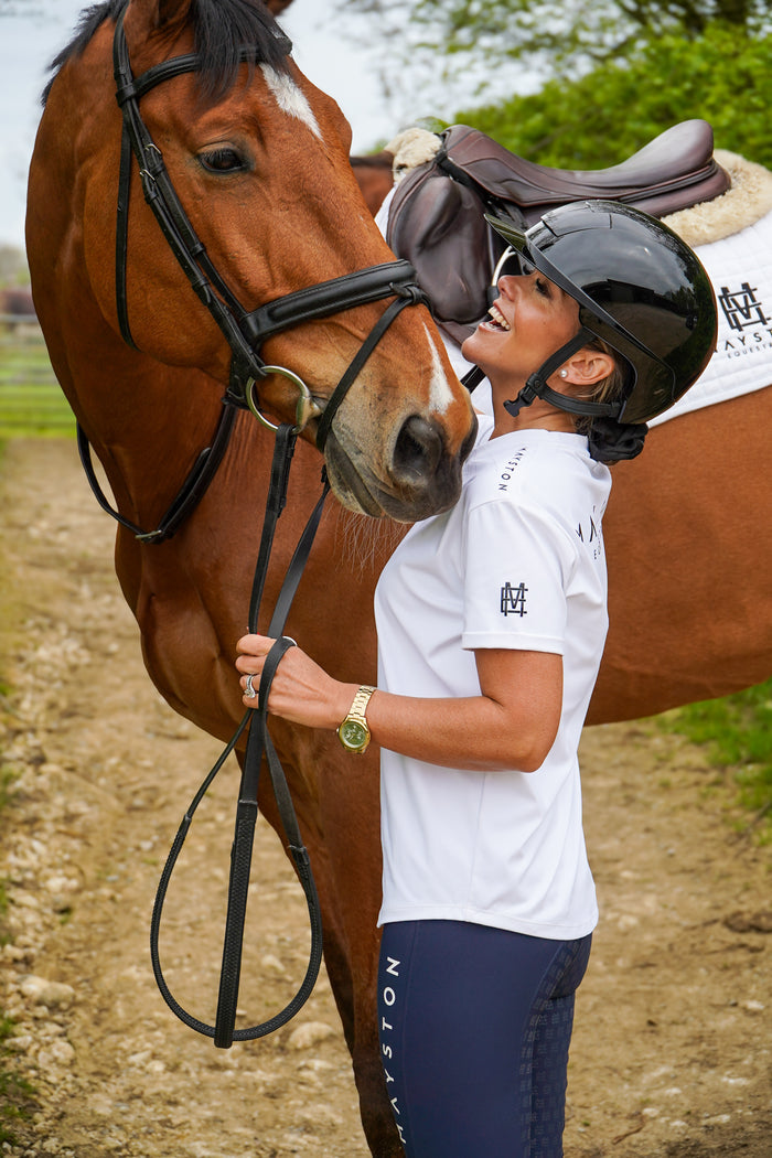 Woman in equestrian attire standing next to a horse on a dirt path. white luxury quality t shirt and fabric is specially crafted for active duty, providing flexibility and ease of movement for any busy lifestyle. An ideal choice for a day at the yard, in the saddle, or during your grooming duties, its loose-fitting design guarantees complete freedom. high gloss black silicone branding