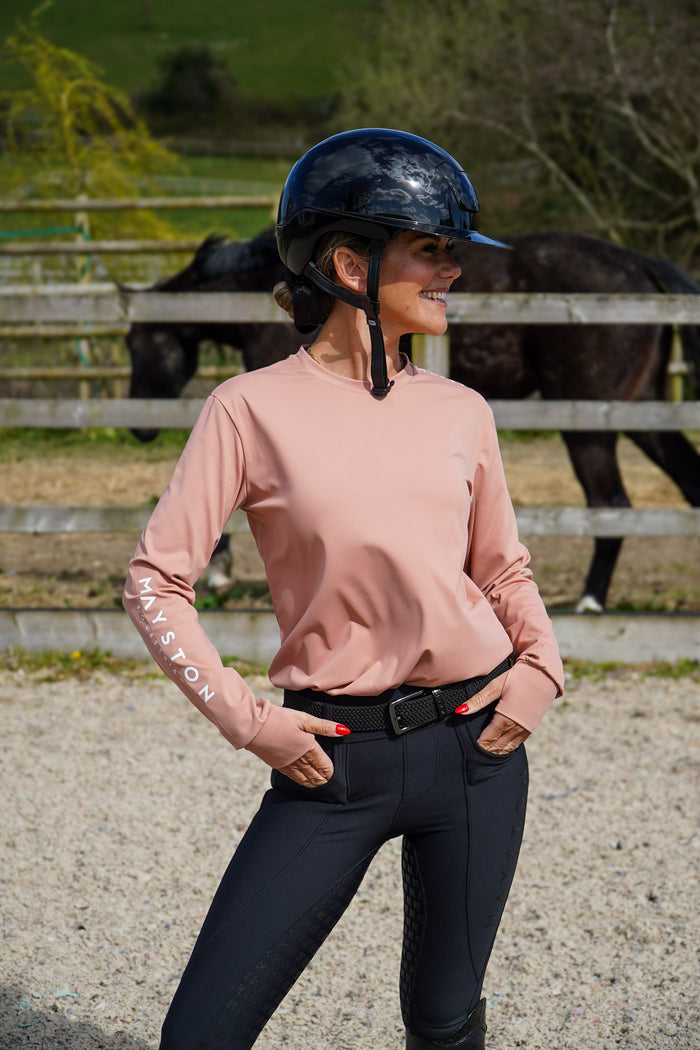 Woman in equestrian attire standing in a stable area with a horse in the background. Crafted from a luxuriously soft technical sports fabric in a breathable nylon blend with stretch, it offers a smooth, lightweight feel that moves effortlessly with you — from stable life to everyday wear. Badminton Blush long sleeve durlston, relaxed fit, not base layer style