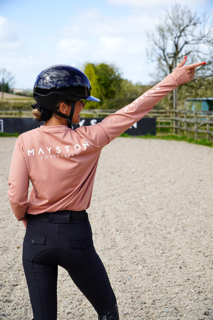 Person wearing a pink 'Mayston' shirt and black riding pants in an equestrian setting. Crafted from a luxuriously soft technical sports fabric in a breathable nylon blend with stretch, it offers a smooth, lightweight feel that moves effortlessly with you — from stable life to everyday wear. Badminton Blush long sleeve durlston, relaxed fit, not base layer style