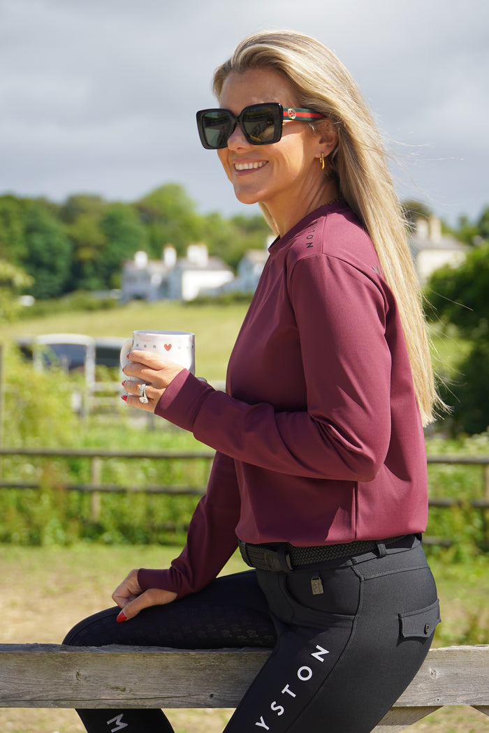 Person in maroon hoodie with sunglasses on, leaning against a wooden post outdoors. burgundy long sleeve durlston sports technical top in buttery-soft nylon sports fabric, this top delivers unbeatable comfort with a sleek, performance feel. From breezy dog walks to competition days, it’s designed to move with you. high gloss silicone branding. thumb holes