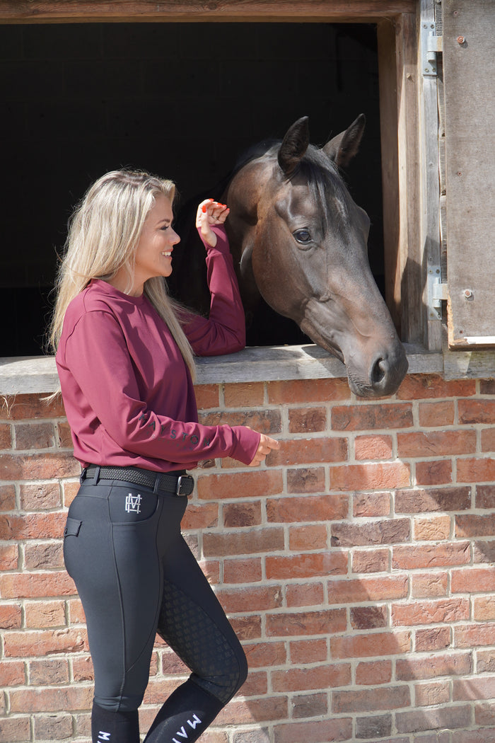 Woman in pink jacket and black breeches interacting with a horse in a stable. burgundy long sleeve durlston sports technical top in buttery-soft nylon sports fabric, this top delivers unbeatable comfort with a sleek, performance feel. From breezy dog walks to competition days, it’s designed to move with you. high gloss silicone branding. thumb holes