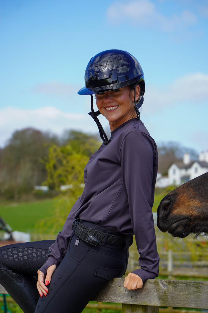 Woman in equestrian attire with a horse in an outdoor setting. grey long sleeve durlston sports technical top in buttery-soft nylon sports fabric, this top delivers unbeatable comfort with a sleek, performance feel. From breezy dog walks to competition days, it’s designed to move with you. high gloss silicone branding. thumb holes