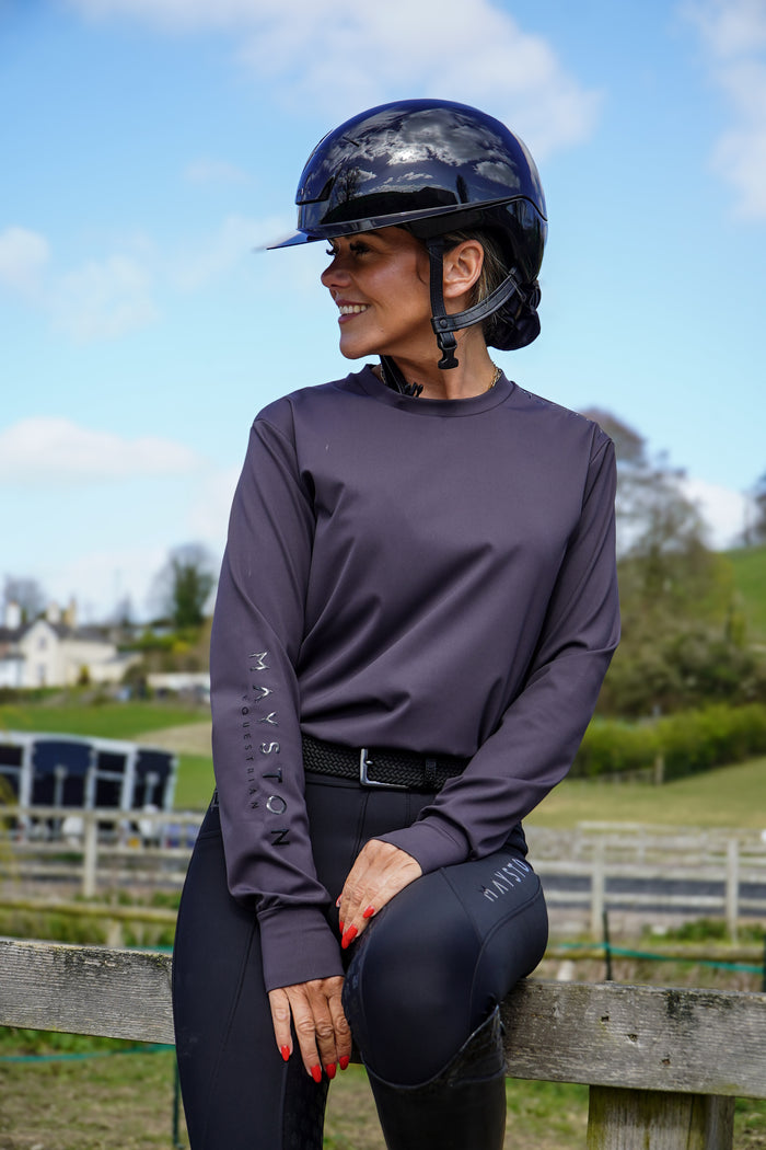Person in equestrian attire with helmet and gloves, sitting on a wooden fence outdoors. grey long sleeve durlston sports technical top in buttery-soft nylon sports fabric, this top delivers unbeatable comfort with a sleek, performance feel. From breezy dog walks to competition days, it’s designed to move with you. high gloss silicone branding. thumb holes