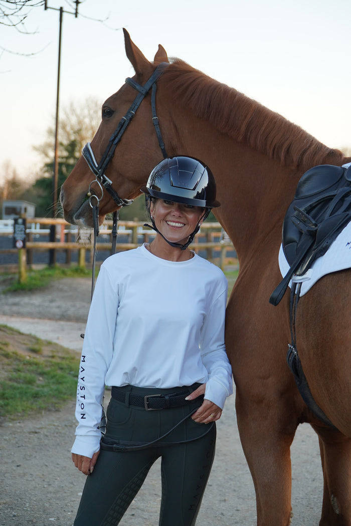 Person riding a horse on a path with buildings in the background. white long sleeve durlston sports technical top in buttery-soft nylon sports fabric, this top delivers unbeatable comfort with a sleek, performance feel. From breezy dog walks to competition days, it’s designed to move with you. high gloss silicone branding. thumb holes