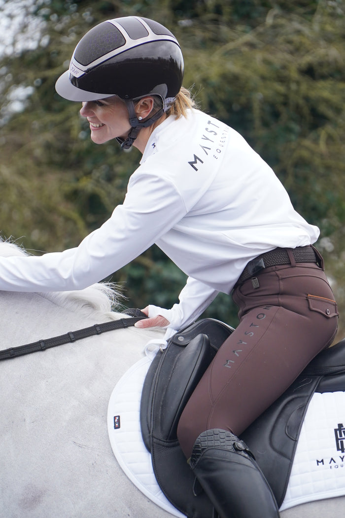 Person adjusting a saddle on a horse with a blurred natural background. white long sleeve durlston sports technical top in buttery-soft nylon sports fabric, this top delivers unbeatable comfort with a sleek, performance feel. From breezy dog walks to competition days, it’s designed to move with you. high gloss silicone branding. thumb holes