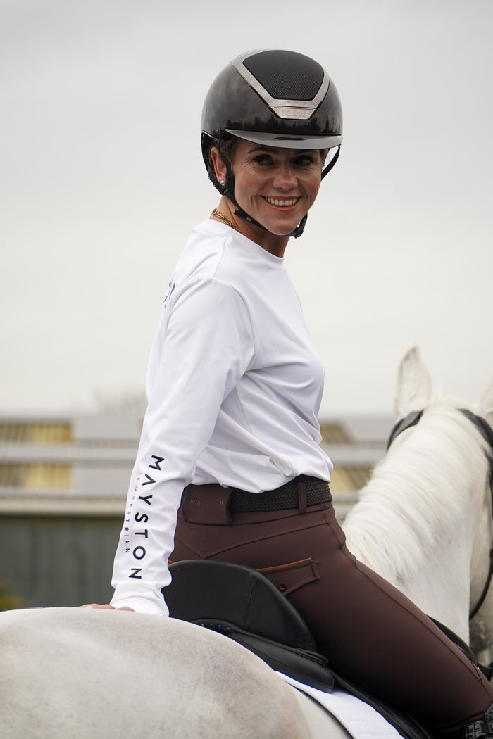 Woman in equestrian attire sitting on a horse with a plain background. white long sleeve durlston sports technical top in buttery-soft nylon sports fabric, this top delivers unbeatable comfort with a sleek, performance feel. From breezy dog walks to competition days, it’s designed to move with you. high gloss silicone branding. thumb holes