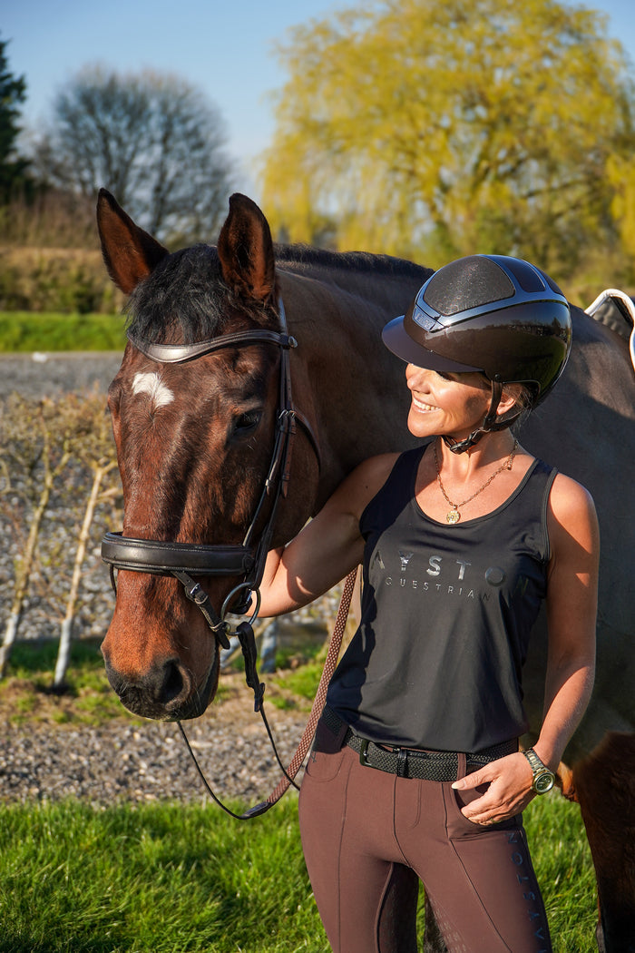 Woman in equestrian gear standing next to a horse in an outdoor setting.  Radipole Racerback Vest, crafted from lightweight, high-quality sports material for effortless comfort and performance. Designed to move with you, it offers breathability and freedom of motion, making it perfect for riding, training, or everyday wear. Finished with bold white high-gloss Mayston silicone branding on the chest and a sleek logo at the rear, this black vest combines sporty style with signature Mayston.