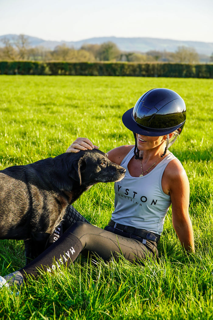 Person sitting on grass with a dog, wearing a helmet and Wiston clothing. Radipole Racerback Vest, crafted from lightweight, high-quality sports material for effortless comfort and performance. Designed to move with you, it offers breathability and freedom of motion, making it perfect for riding, training, or everyday wear. Finished with bold high-gloss Mayston silicone branding on the chest and a sleek logo at the rear, this soft grey vest combines sporty style with signature Mayston.