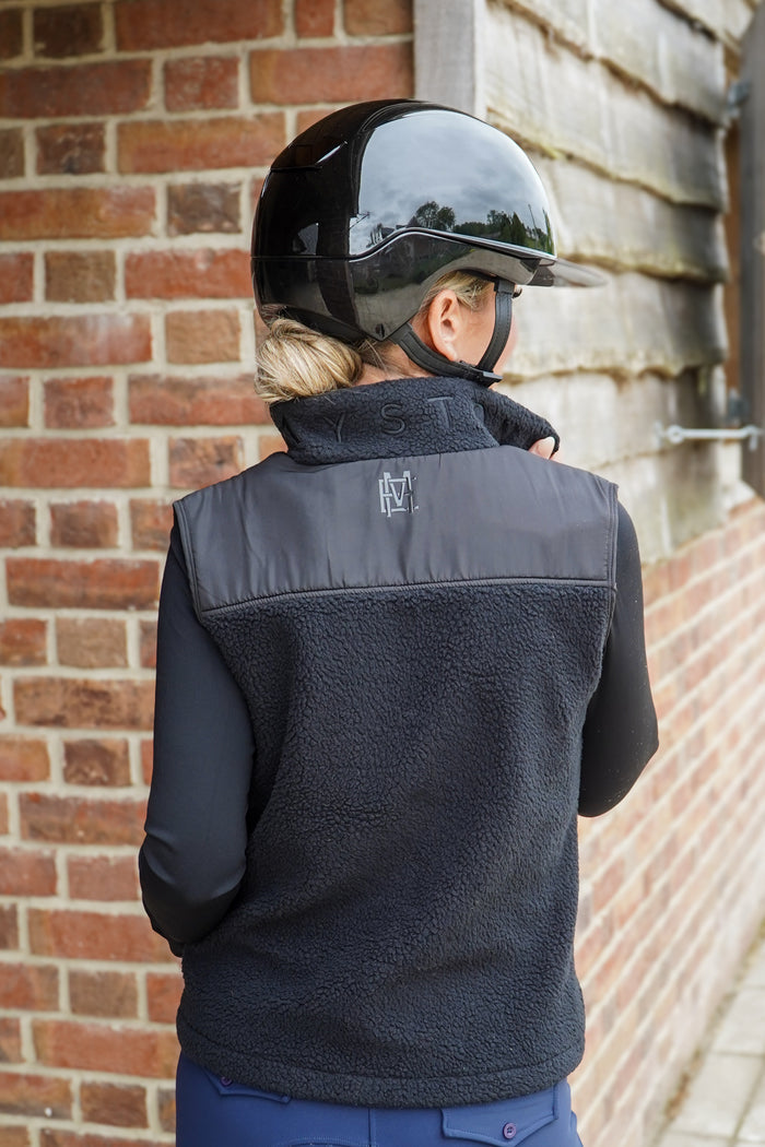 Person wearing a black equestrian vest with a visible brand logo, standing against a brick wall. MAYSTON Tolpuddle Teddy Gilet in Black. Crafted from plush cotton-polyester blend, it delivers warmth without bulk, leaving your arms free for riding, yard work. Colour-matched MAYSTON embroidery on the collar and a discreet silicone logo keep it refined and effortlessly on-brand. Stretch-lined interior and double zip ensures a flattering fit.