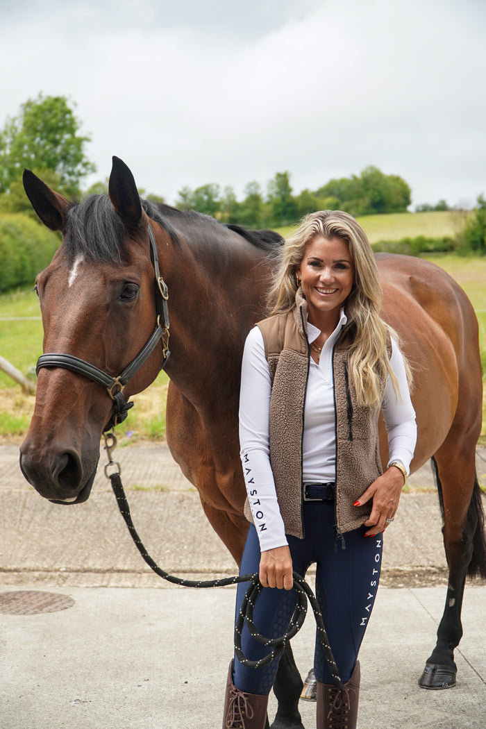 Woman standing next to a horse in an outdoor setting.  Luxury teddy Fleece Gilet Waistcoat. Quality embroidery. multiple pockets. Subtle branding, elegant classic design. Stretch lining