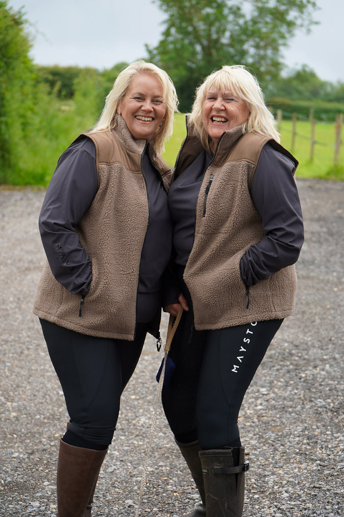 Two women wearing matching brown vests and dark pants standing outdoors on a gravel path with greenery in the background.  Luxury teddy Fleece Gilet Waistcoat. Quality embroidery. multiple pockets. Subtle branding, elegant classic design. Stretch lining