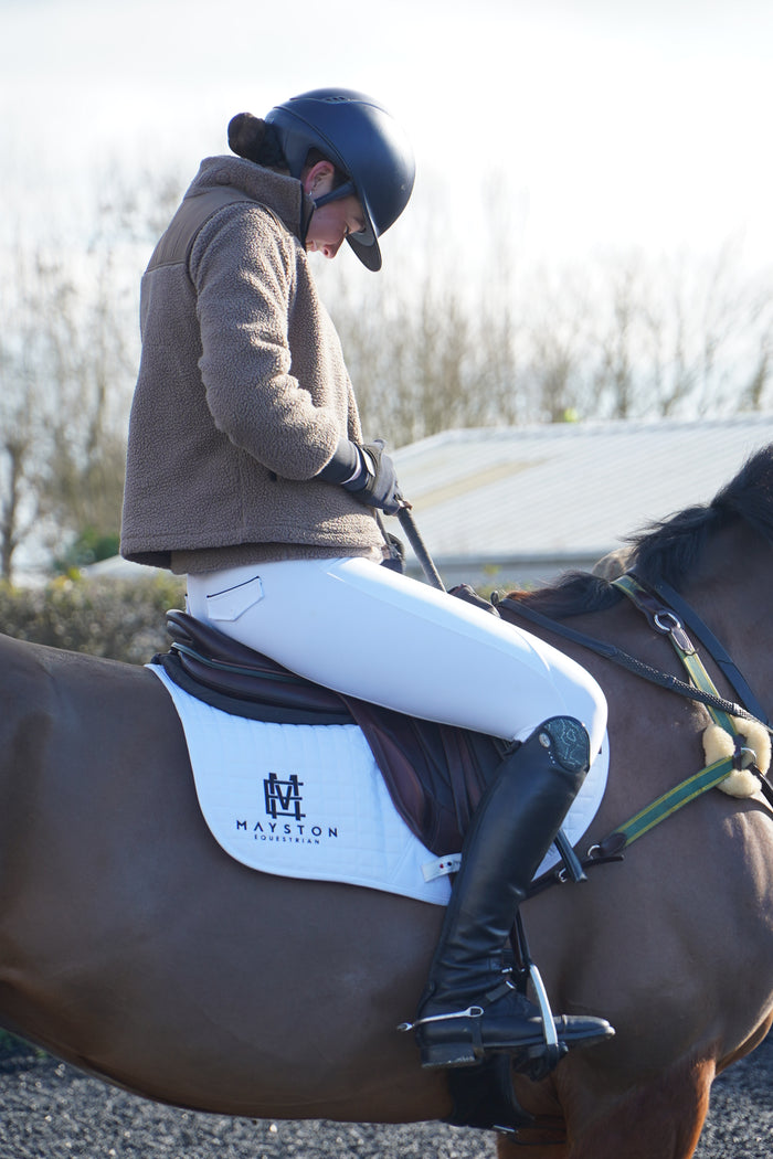 Person riding a horse with a saddle and bridle, standing by a body of water. Premium white competition breeches with a Full Silicone seat. Stretch high waisted Jodhpurs. Deep pockets, supportive, comfortable and compression quality fabric. Elegant detailing with subtle branding.