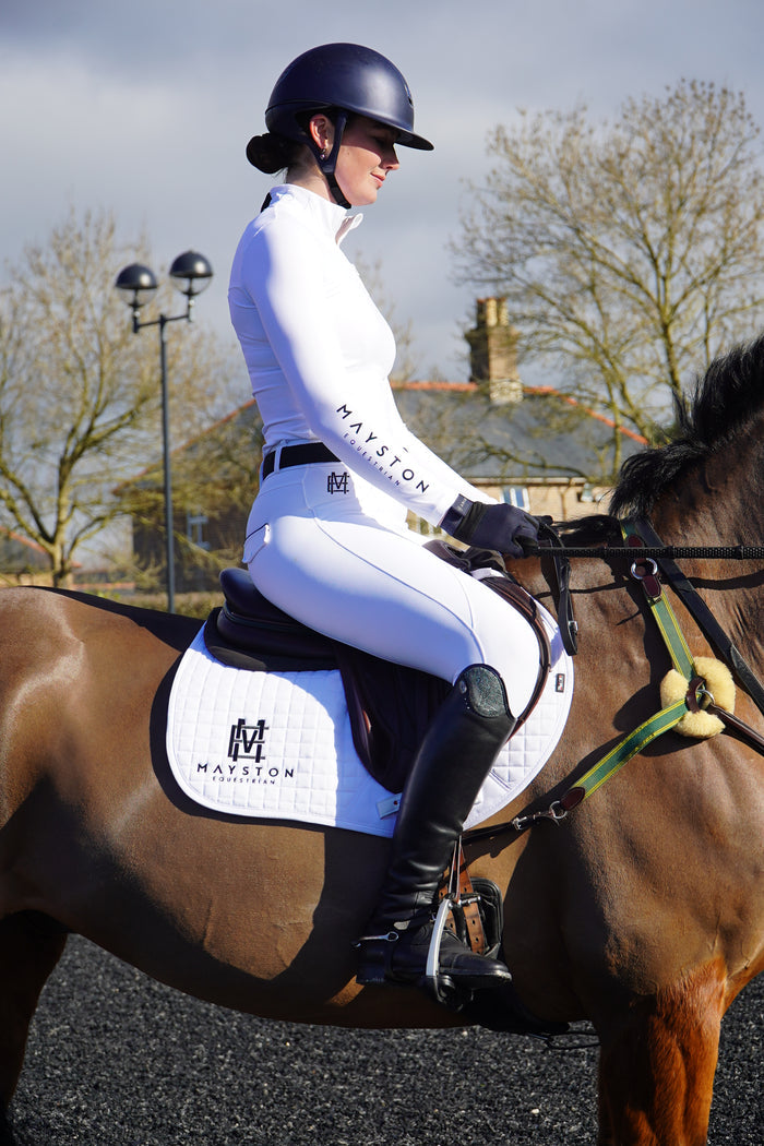 Person riding a horse with a blurred background. Sleek silhouette baselayer showshirt top subtle Mayston branding, maximum coverage without compromising style,  next-level performance with a bold, minimalist look. Moisture-wicking, breathable fabric with four-way stretch, it keeps you cool and comfortable. Training, Everyday or competition wear. long sleeve Sleeve 1/4 zip with standup collar. Suble Black Mayston branding.